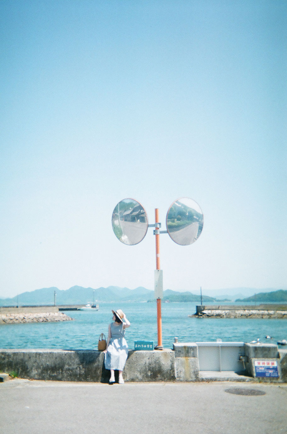 青空と海を背景に岸壁に座る女性と、カーブミラーの写真